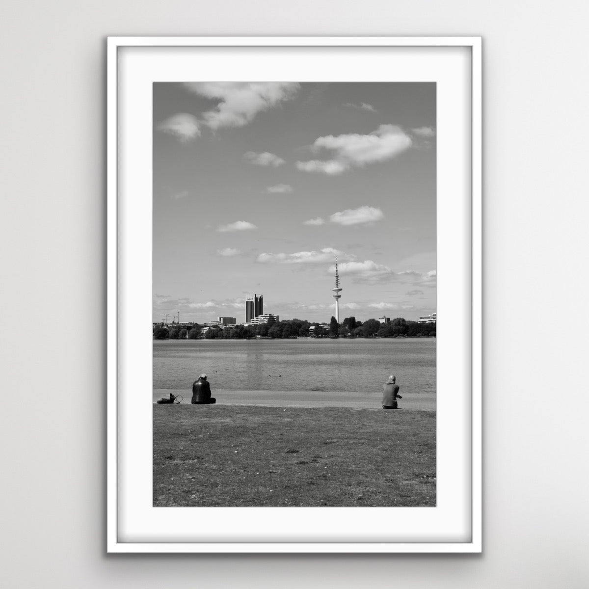 Schwarz-Weiss-Fotografie eines ruhigen Sees mit einer grossen Rasenfläche im Vordergrund. Zwei Personen sitzen auf der Wiese und blicken in Richtung Wasser. Am gegenüberliegenden Ufer sind mehrere Gebäude und ein hoher Fernsehturm zu sehen. Der Himmel ist teils bewölkt.