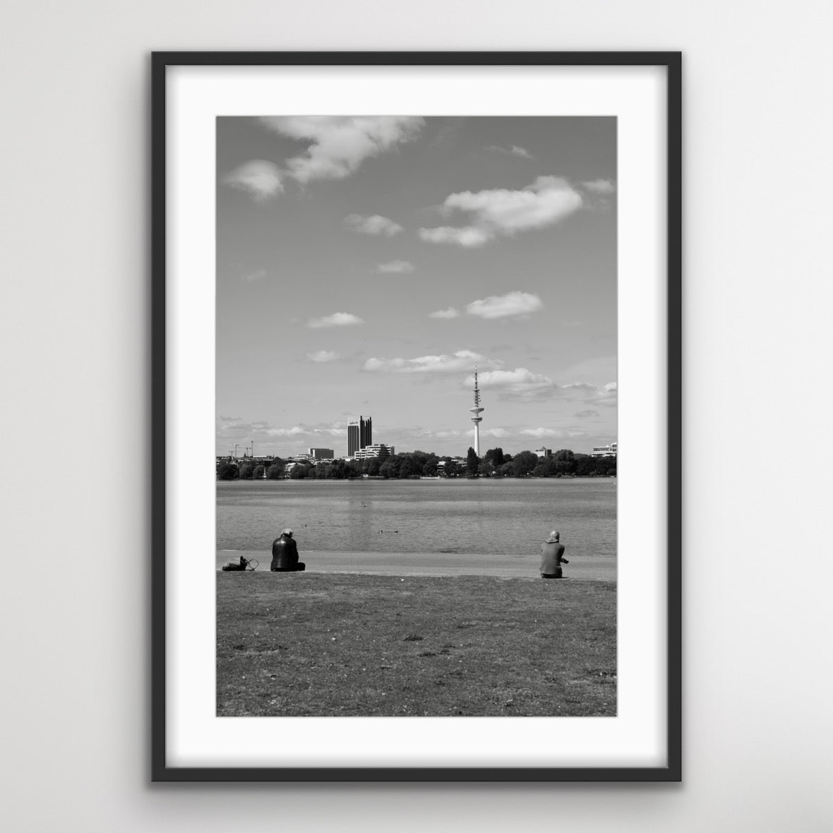 Schwarz-Weiss-Fotografie eines ruhigen Sees mit einer grossen Rasenfläche im Vordergrund. Zwei Personen sitzen auf der Wiese und blicken in Richtung Wasser. Am gegenüberliegenden Ufer sind mehrere Gebäude und ein hoher Fernsehturm zu sehen. Der Himmel ist teils bewölkt.