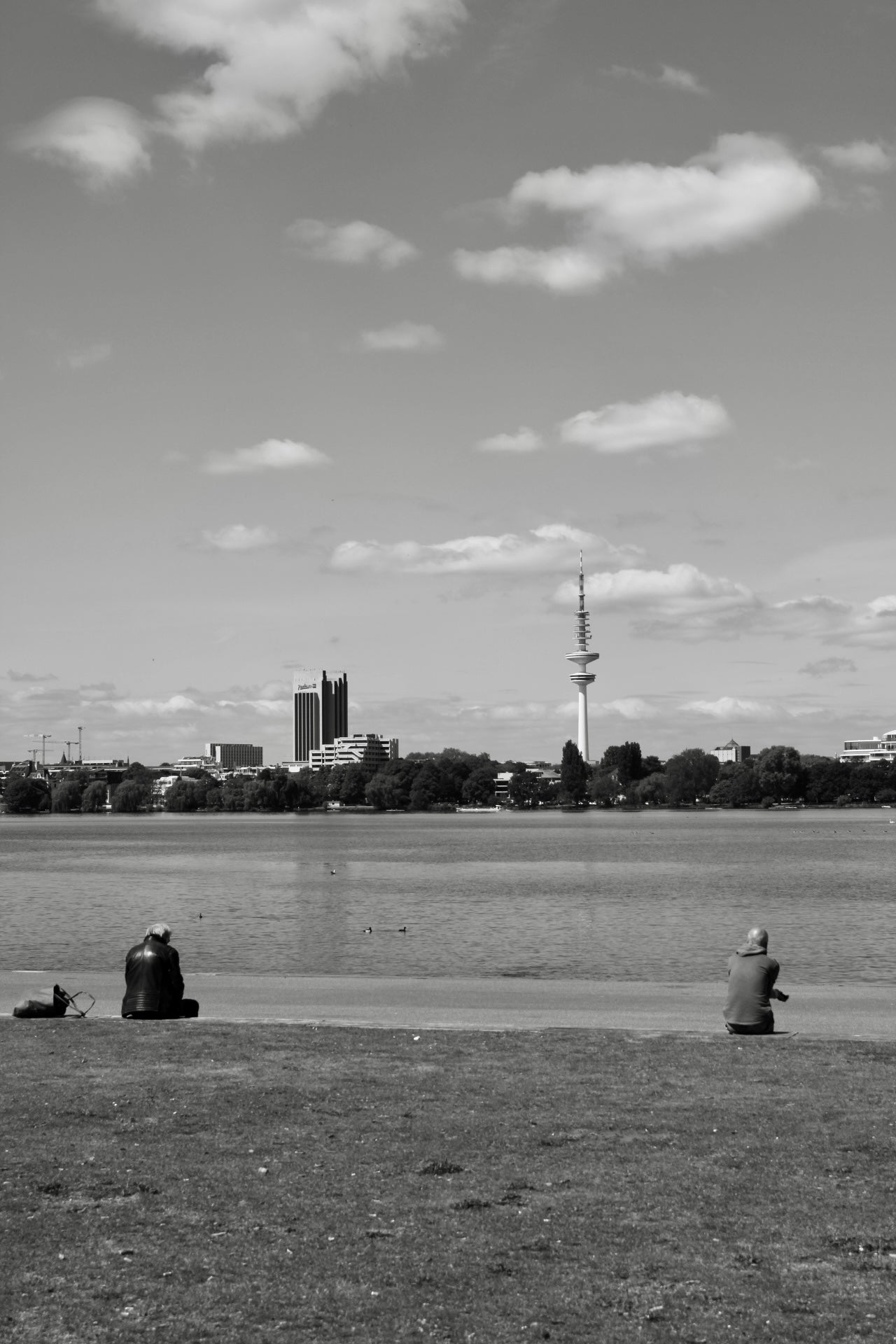 Schwarz-Weiss-Foto eines ruhigen Sees mit einer Rasenfläche im Vordergrund. Zwei Personen sitzen am Ufer und schauen auf das Wasser. Auf dem See schwimmen einige Enten. Am gegenüberliegenden Ufer stehen mehrere Gebäude und ein hoher Fernsehturm, der sich in die Wolken am Himmel erhebt.