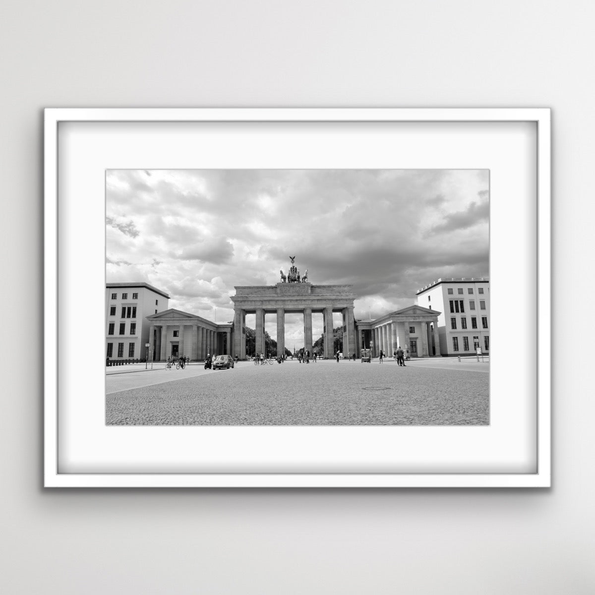 Schwarz-Weiss-Fotografie des Brandenburger Tors in Berlin. Das historische Tor befindet sich in der Mitte des Bildes, flankiert von zwei Nebengebäuden. Mehrere Menschen spazieren oder fahren mit Fahrrädern über den grossen, gepflasterten Platz vor dem Tor. Im Hintergrund ist ein bewölkter Himmel zu sehen.