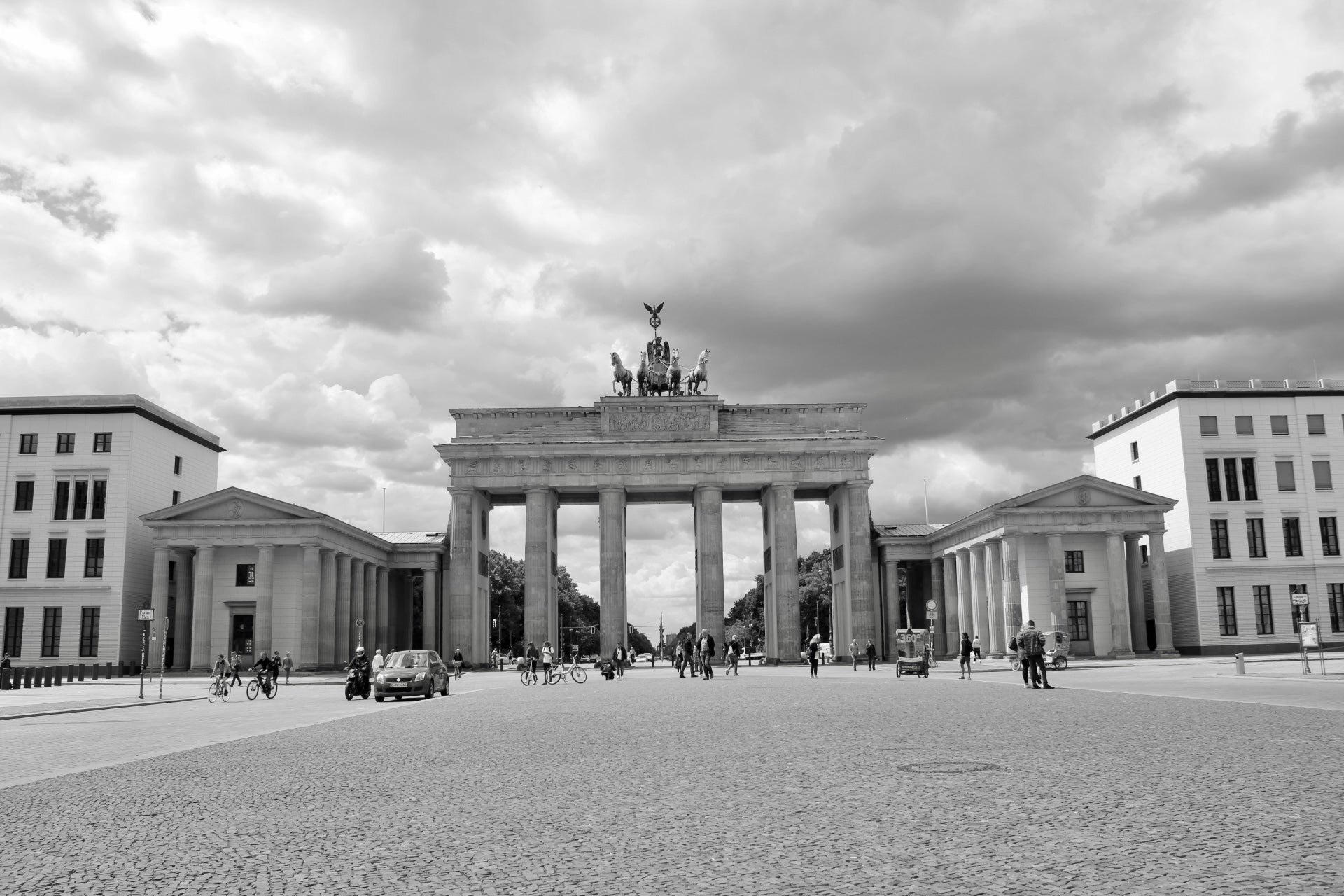 Schwarz-Weiss-Foto des Brandenburger Tors in Berlin. Vor dem Tor befinden sich mehrere Personen, die spazieren oder mit dem Fahrrad fahren. Einige Autos und Motorräder sind ebenfalls auf dem Platz zu sehen. Auf dem Brandenburger Tor steht die Quadriga-Statue, und im Hintergrund ist ein dichter, bewölkter Himmel zu erkennen.