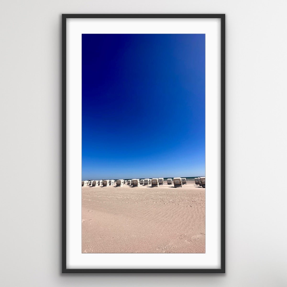 Gerahmtes Foto von einem Strand mit mehreren Strandkörben in einer Reihe. Der Strand ist menschenleer, und der Himmel darüber ist wolkenlos und tiefblau. Die Aufnahme zeigt den weiten, sandigen Strandbereich im Vordergrund und die am Horizont angeordneten Strandkörbe, die auf das Meer ausgerichtet sind.