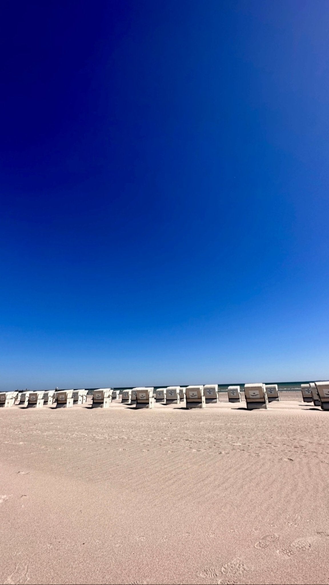 Strandaufnahme mit einer Reihe von weissen Strandkörben am Horizont. Der Vordergrund zeigt einen weiten, unberührten Sandstrand. Darüber erstreckt sich ein klarer, tiefblauer Himmel ohne Wolken.