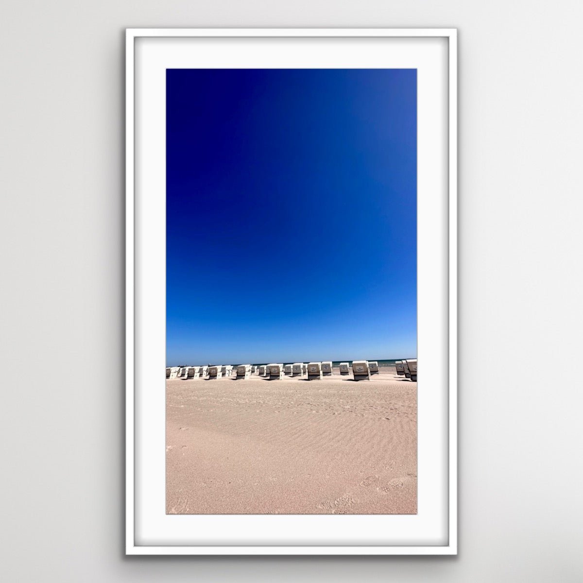 Gerahmtes Foto von einem Strand mit mehreren Strandkörben in einer Reihe. Der Strand ist menschenleer, und der Himmel darüber ist wolkenlos und tiefblau. Die Aufnahme zeigt den weiten, sandigen Strandbereich im Vordergrund und die am Horizont angeordneten Strandkörbe, die auf das Meer ausgerichtet sind.